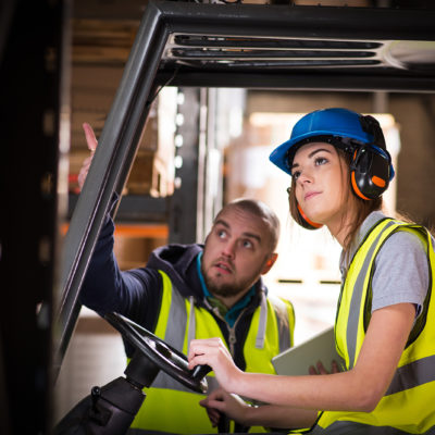 A young female forklift driver is being trained on approaching the racking of the warehouse . A male colleague or trainer can be seen advising her on how to perform the manoeuvre . She is wearing a hi vis jacket , hard hat and ear defenders .