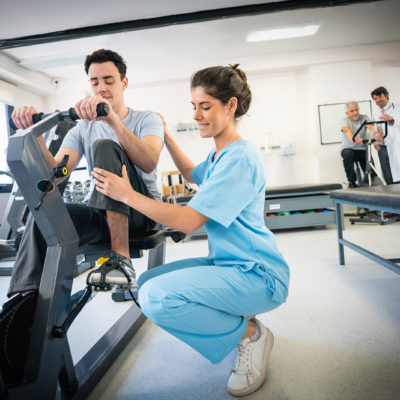 Physiotherapist correcting the posture of a patient on a static bicycle while doctor and senior patient are working out at the background on a static bicycle